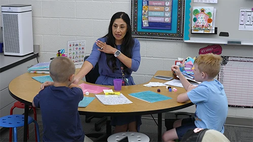 Teacher instructing two young students at a round classroom table