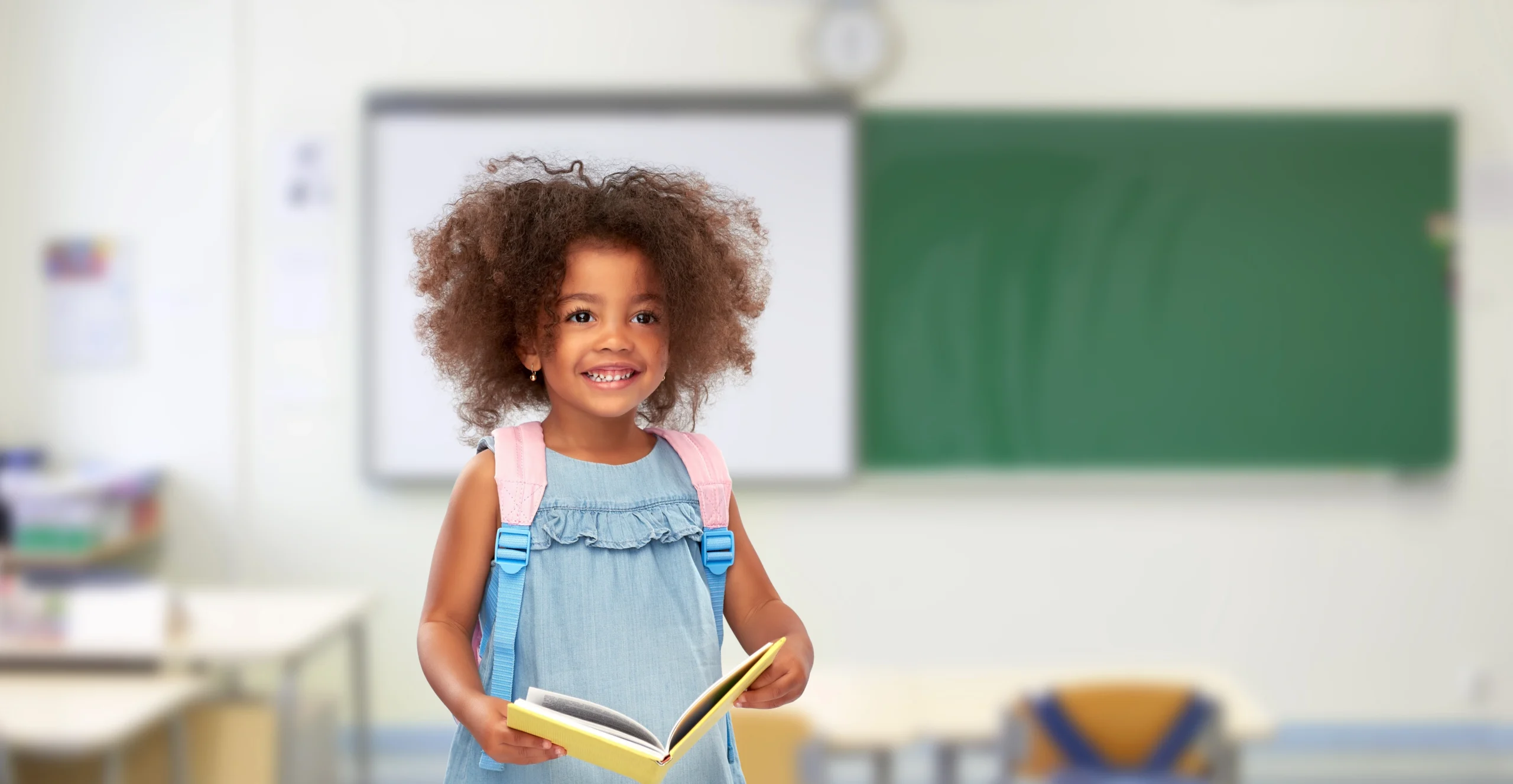 Smiling young girl with backpack holding book in blurred classroom