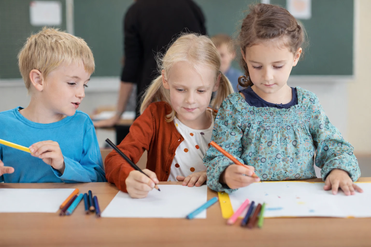 Three school children sitting at a desk drawing with colored pencils